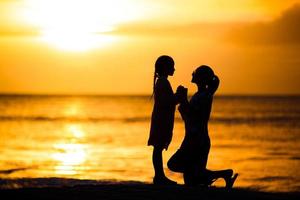 weinig meisje en gelukkig moeder silhouet in de zonsondergang Bij de strand foto