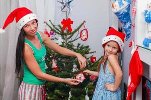 jong moeder en haar aanbiddelijk weinig meisje in de kerstman claus hoeden versieren hun Kerstmis boom foto