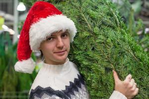 portret van jong Mens in de kerstman hoed buying Kerstmis boom en en tonen duimen omhoog foto
