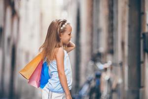 portret van aanbiddelijk weinig meisje wandelen met boodschappen doen Tassen buitenshuis in Europese stad. foto