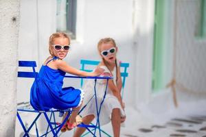 twee meisjes in blauw jurken zittend Aan blauw stoelen en tafel Aan straat van typisch Grieks traditioneel dorp met wit huizen Aan Mykonos eiland, Griekenland, Europa. foto