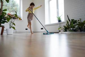 een meisje dansen met een dweilen naar schoon de verdieping in een nieuw huis - algemeen schoonmaak in een leeg kamer, de vreugde van in beweging, helpen met de huiswerk foto