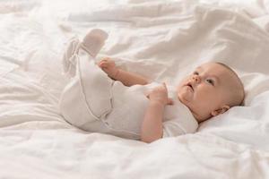 portret van een schattig baby in een wit bodysuit Aan een bed Bij huis met wit linnengoed. pasgeboren baby Bij huis, gelukkig moederschap. hoog kwaliteit foto