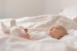 portret van een schattig baby in een wit bodysuit Aan een bed Bij huis met wit linnengoed. pasgeboren baby Bij huis, gelukkig moederschap. hoog kwaliteit foto