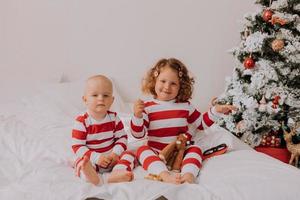 kinderen in rood en wit pyjama eten Kerstmis snoepgoed zittend in bed. broer en zus, jongen en meisje delen geschenken. Kerstmis ochtend. levensstijl. ruimte voor tekst. hoog kwaliteit foto