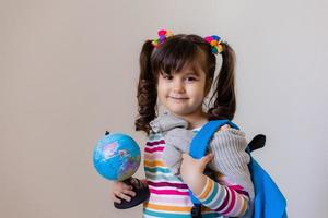 een gelukkig weinig peuter- meisje met een rugzak en een wereldbol Aan een licht achtergrond, ruimte voor tekst. peuter- opleiding, op reis met kinderen. hoog kwaliteit foto