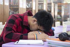 jong Aziatisch jongen draagt regenboog polsbandje, zittend in bibliotheek en nemen een dutje Aan tafel terwijl aan het doen zijn moeilijk school- project werk, concept voor verhogen tieners en lgbt mensen dagelijks leven werkzaamheid. foto