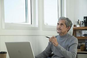 portret ouderen senior Aziatisch Mens zit in de buurt glas venster in de ochtend- naar werk van huis en controle zijn bedrijf Aan zijn laptop Aan tafel ernstig, zacht en selectief focus. foto