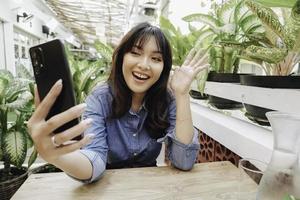 een gelukkig Aziatisch vrouw in een restaurant, vervelend een blauw overhemd en Holding haar telefoon foto