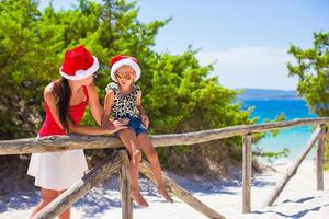 jong moeder en weinig meisje in rood Kerstmis hoed Bij tropisch strand foto