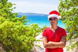 jong Mens in rood Kerstmis hoed Aan tropisch strand in de buurt de sparren foto