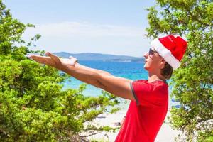 jong Mens in rood Kerstmis hoed Aan tropisch strand in de buurt de sparren foto