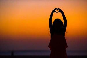 silhouet van weinig meisje maken hart Bij zonsondergang Aan de strand foto