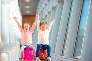 aanbiddelijk weinig meisjes hebben pret in luchthaven zittend Aan koffer aan het wachten voor vlucht foto