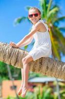 aanbiddelijk weinig meisje zittend Aan palm boom gedurende zomer vakantie Aan wit strand foto