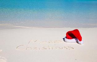 de kerstman hoed Aan wit zanderig strand en vrolijk Kerstmis geschreven in de zand foto