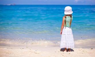 achterzijde visie van weinig meisje in hoed op zoek Bij de zee Aan wit zand strand foto