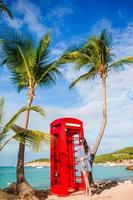 mooi vrouw in de buurt rood telefoon stand in van Dickenson baai antigua. mooi landschap met een klassiek telefoon stand Aan de wit zanderig strand in antigua foto