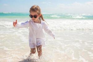 schattig kleuter meisje Bij wit jurk staand in Ondiep water Aan exotisch strand foto