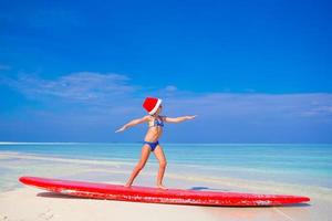 aanbiddelijk weinig meisje in de kerstman hoed praktijk surfing positie Aan surfboard gedurende vakantie foto