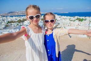 twee meisjes in blauw jurken nemen selfie foto buitenshuis. kinderen achtergrond van typisch Grieks traditioneel dorp met wit muren en kleurrijk deuren Aan Mykonos eiland, in Griekenland