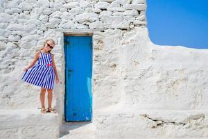 schattig meisje in blauw jurk hebben pret buitenshuis in de buurt paraportiani kerk. kind Bij straat van typisch Grieks traditioneel dorp met wit muren en kleurrijk deuren Aan Mykonos eiland, in Griekenland foto