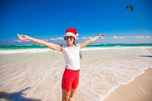 jong vrouw in de kerstman hoed wandelen verspreiding haar handen Aan wit zanderig strand foto