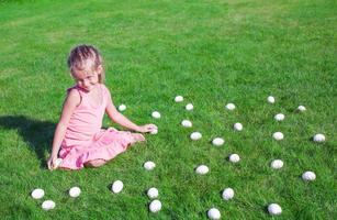 weinig meisje met wit Pasen eieren in de werf Aan groen gras foto
