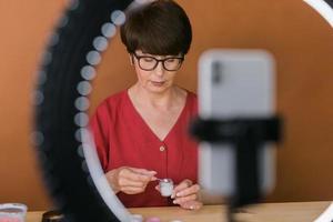 middelbare leeftijd vrouw schoonheid blogger of influencer met bedenken Product opname video in kamer Bij huis. gebruik makend van ring lamp en smartphone foto