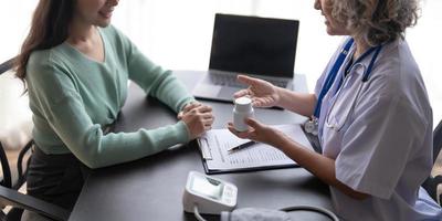 vrouw senior dokter is lezing medisch geschiedenis van vrouw geduldig en sprekend met haar gedurende overleg in een Gezondheid kliniek. arts in laboratorium jas zittend achter een laptop in ziekenhuis kantoor. foto