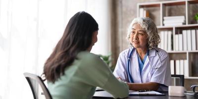 vrouw senior dokter is lezing medisch geschiedenis van vrouw geduldig en sprekend met haar gedurende overleg in een Gezondheid kliniek. arts in laboratorium jas zittend achter een laptop in ziekenhuis kantoor. foto