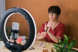 middelbare leeftijd vrouw schoonheid blogger of influencer met bedenken Product opname video in kamer Bij huis. gebruik makend van ring lamp en smartphone foto