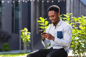 vrolijk Afrikaanse Amerikaans vent gebruik makend van mobiel telefoon zittend Aan bank in stad park buitenshuis