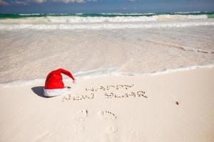 de kerstman hoed Aan wit zanderig strand en gelukkig nieuw jaar geschreven in de zand foto