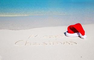 vrolijk Kerstmis geschreven Aan tropisch strand wit zand met Kerstmis hoed foto
