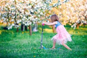 weinig meisje hebben pret Aan scooter in bloeiend appel boom tuin Aan voorjaar dag foto