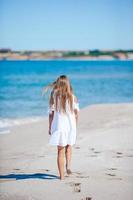 terug visie van aanbiddelijk weinig meisje met lang haar- in wit jurk wandelen Aan tropisch strand vakantie foto