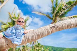 aanbiddelijk weinig meisje zittend Aan palm boom gedurende zomer vakantie Aan wit strand foto