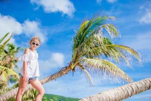 aanbiddelijk weinig meisje zittend Aan palm boom gedurende zomer vakantie Aan wit strand foto