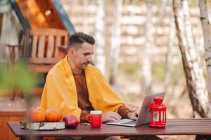 gelukkig jong Mens werken Aan laptop en drinken koffie zittend Bij de houten tafel buitenshuis foto