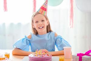 Kaukasisch meisje is dromerig glimlachen en op zoek Bij verjaardag regenboog taart. feestelijk kleurrijk achtergrond met ballonnen. verjaardag partij en wensen concept. foto