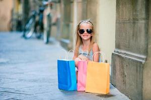 portret van aanbiddelijk weinig meisje wandelen met boodschappen doen Tassen buitenshuis in Europese stad. mode kleuter kind in Italiaans stad met haar boodschappen doen foto