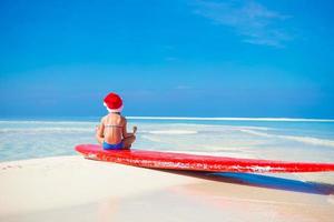 weinig schattig meisje in de kerstman hoed Aan wit strand gedurende vakantie in lotus positie foto