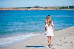 terug visie van aanbiddelijk weinig meisje met lang haar- in wit jurk wandelen Aan tropisch strand vakantie foto