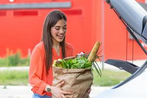 jong vrouw rijden boodschappen doen kar vol van voedsel Aan de buitenshuis parkeren. jong vrouw in auto park, bezig met laden boodschappen doen in bagageruimte van auto. boodschappen doen met succes gedaan. vrouw zetten Tassen in auto na boodschappen doen foto