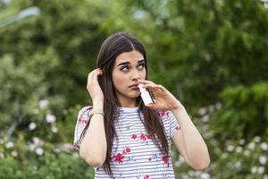 stuifmeel allergie concept. jong vrouw gebruik makend van neus- verstuiven voor haar stuifmeel en gras allergieën. bloeiend bomen in achtergrond. voorjaar seizoensgebonden allergieën en Gezondheid problemen. foto