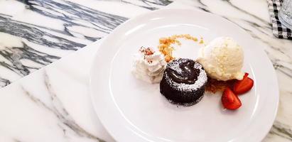 vlak leggen of top visie van chocola lava taart met vanille ijs room, geslagen room en gesneden van aardbei in bord Aan wit marmeren tafel. toetje en zoet voedsel concept foto