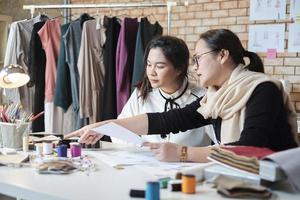 Aziatisch middelbare leeftijd vrouw mode ontwerper geeft les jong tiener stagiair kleermaker in studio, tekening schetsen stijl, en kleding stof naaien voor jurk ontwerp verzameling ideeën, professioneel winkel klein bedrijf. foto