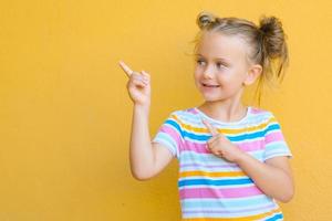 glimlachen weinig kind meisje is aangetrokken door aandacht richten vinger Bij kopiëren ruimte. de concept van reclame producten , goederen en Diensten. geïsoleerd Aan geel achtergrond foto