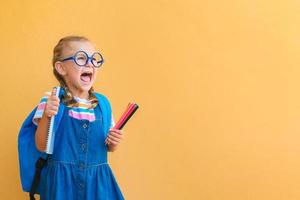 emotioneel opgewonden verrassing gezicht schoolmeisje kind in bril in school- uniform houdt gekleurde potloden, notitieboekje, rugzak Open mond naar leeg kopiëren ruimte attent geïsoleerd Aan geel achtergrond foto